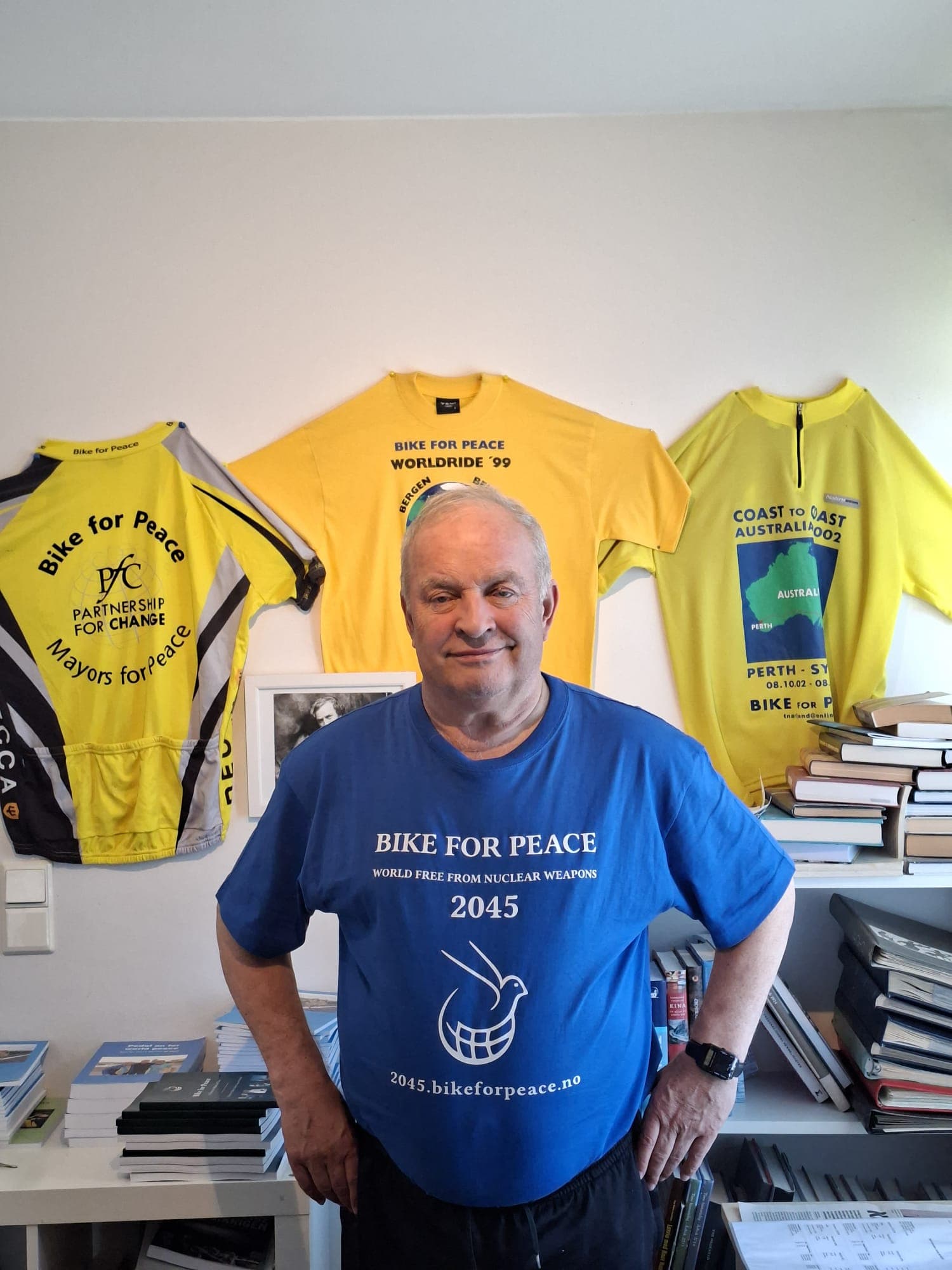 Tore Naerland wearing the blue anniversary shirt. The print on the front reads Bike for Peace, World free from nuclear weapons, 2045, above an emblem of a dove in a globe, and the web address 2045.bikeforpeace.no. Three older Bike for Peace jerseys hang on the wall behind him.